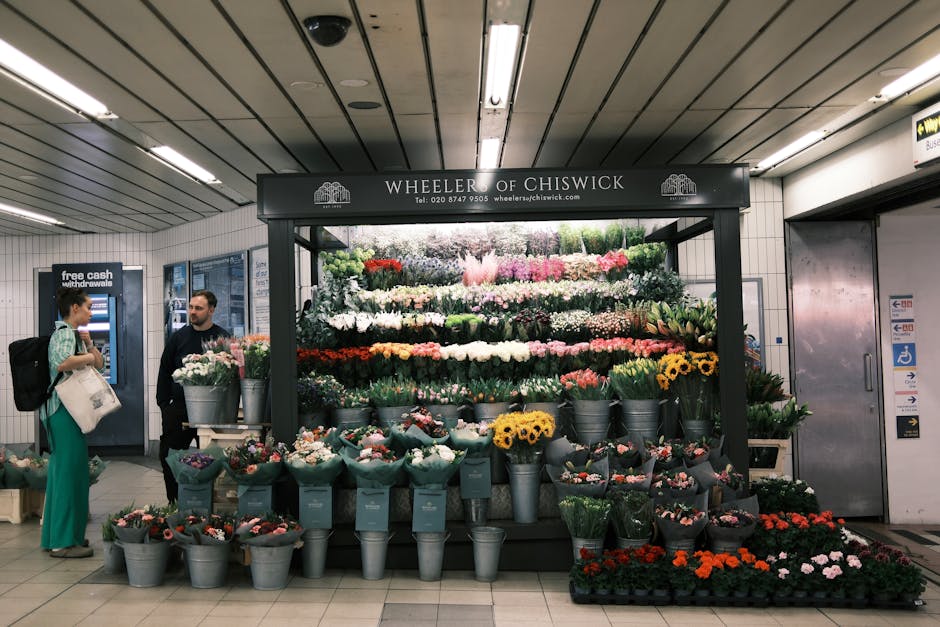 Inside a subway station, a flower stall named 'Wheeler of Chiswick' displays an array of potted flowers and plants arranged on tiered shelves. The flowers include various colours such as white, pink, red, yellow, and purple, with some in metal buckets and others wrapped in paper. Behind the stall, the station’s ceiling features metallic panels with linear lighting fixtures, illuminating the scene evenly. To the left, a woman with dark hair tied back, dressed in a teal outfit and carrying a backpack, stands facing a man who is wearing dark clothing, engaging in conversation. Adjacent to them, a self-service cash machine with a blue interface is mounted on the tiled wall, which is lined with informational posters and signs, including accessibility notices. The station's environment appears clean and well-maintained, with a neutral-toned floor and a partially visible elevator door to the right. The overall scene captures a moment of private, on-site retail activity within an urban underground transportation setting, aligned with alternative waste and rubbish removal context through the visible organized display and station infrastructure.