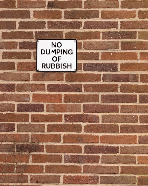 A rectangular black and white metal sign mounted on a red brick wall displaying the message 'NO DUMPING OF RUBBISH' in bold black letters. The wall features uniform, horizontally laid bricks with varying shades of red and brown, and light mortar joints between them. The sign is positioned near the upper left of the image, with the brickwork continuing in a regular pattern across the background. The lighting suggests natural daylight, casting soft shadows that highlight the texture of the bricks and the sign. The scene reflects a controlled environment where private or municipal waste disposal might be regulated, aligning indirectly with rubbish removal and alternative waste handling services provided by companies such as Rubbish Removal Chiswick.