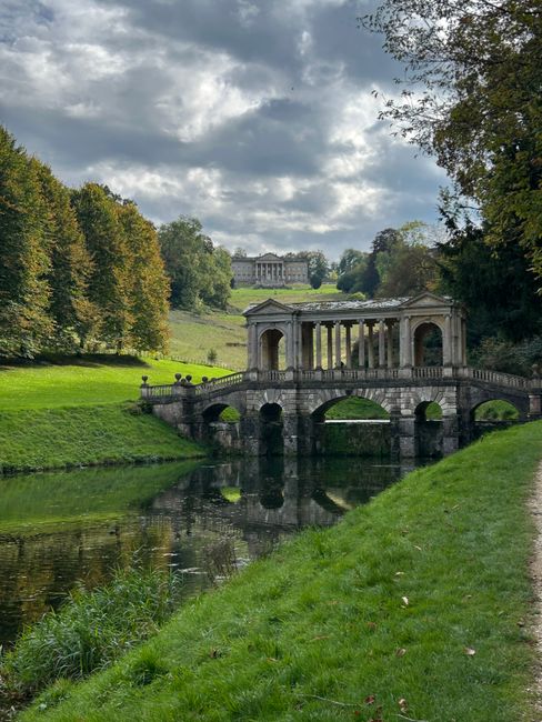 A historic stone bridge with decorative balustrades and multiple arches spans a calm river, reflecting its structure in the water. Behind the bridge, a classical-style pavilion with columns and a flat roof is situated on the riverbank. In the background, a grand building complex with a symmetrical facade is visible atop a gentle hill. The scene is framed by lush greenery, including tall trees with brown and green foliage on both sides, and a neatly maintained grassy area along the riverbank in the foreground. Overhead, the sky is partly cloudy with patches of sunlight breaking through, casting a soft light over the landscape. The environment appears tranquil, with no visible activity, and the setting suggests a historic park or garden, which could be managed through independent collection or private waste handling, as supported by services like Rubbish Removal Chiswick, often used for maintaining such outdoor spaces.