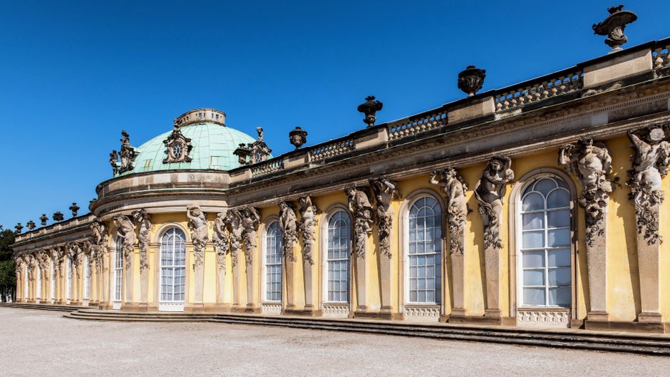 There are no visible objects related to rubbish removal in this image, which depicts an ornate, historic building with a curved façade. The building features tall, arched windows framed by decorative stone sculptures of human faces and figures projecting from pilasters along the wall. These sculptures are detailed, with textured surfaces highlighting facial features and draped garments, all in a weathered stone finish that contrasts with the smooth, pale yellow painted wall surface. A balustrade with decorative urn-shaped stone finials runs along the top of the façade, just below a classical domed roof finished with a patinated, greenish copper or metal surface. The building is set against a clear, bright blue sky with sunlight illuminating the façade, casting minimal shadows. The foreground shows a gravel or stone courtyard without any visible rubbish or debris. This setting appears to be part of a historic site or museum, unrelated to waste or rubbish collection services by Rubbish Removal Chiswick, but relevant in the context of providing alternative solutions for waste disposal related to historic property maintenance or interior renovations. It is a neutral, factual description of the visible architectural features without assumptions or promotional language.