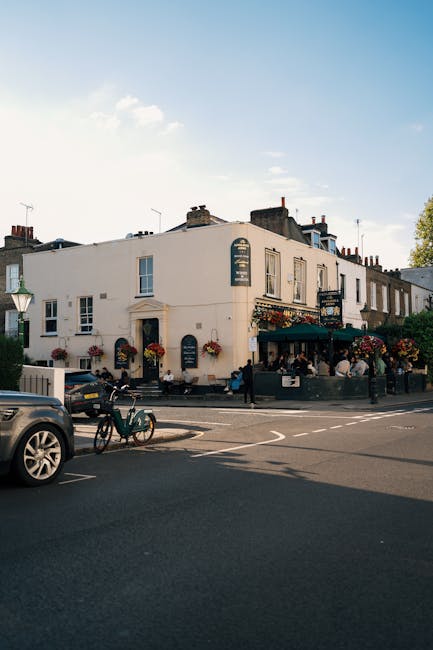 A street scene featuring a white, two-storey building with multiple windows and a curved corner façade, which houses a pub or café indicated by outdoor seating and hanging flower baskets. Several customers sit under a green awning on the right side of the building, with some standing nearby. In the foreground, there is a dark grey car parked partially on the road and a green bicycle resting against the curb, suggesting local non-kerbside waste management or independent collection services may be involved in rubbish removal for the area. The street is lined with black-and-white road markings, and a traditional lamppost is visible on the left. The background shows a clear sky with minimal cloud cover, and neighbouring buildings are partly visible behind the main structure. The scene offers a typical urban environment with a focus on pedestrian activity and local businesses, which may generate waste requiring professional rubbish removal services.