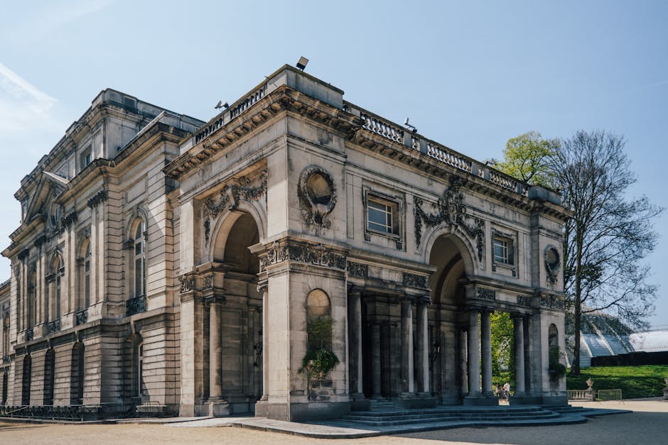 There are no visible objects related to rubbish removal in this image, which depicts an ornate, historic building with a curved façade. The building features tall, arched windows framed by decorative stone sculptures of human faces and figures projecting from pilasters along the wall. These sculptures are detailed, with textured surfaces highlighting facial features and draped garments, all in a weathered stone finish that contrasts with the smooth, pale yellow painted wall surface. A balustrade with decorative urn-shaped stone finials runs along the top of the façade, just below a classical domed roof finished with a patinated, greenish copper or metal surface. The building is set against a clear, bright blue sky with sunlight illuminating the façade, casting minimal shadows. The foreground shows a gravel or stone courtyard without any visible rubbish or debris. This setting appears to be part of a historic site or museum, unrelated to waste or rubbish collection services by Rubbish Removal Chiswick, but relevant in the context of providing alternative solutions for waste disposal related to historic property maintenance or interior renovations. It is a neutral, factual description of the visible architectural features without assumptions or promotional language.