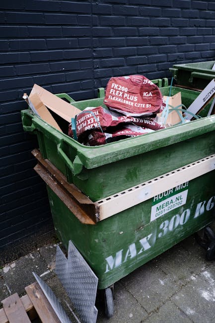 A large green wheeled waste bin filled with black and red packaging material, positioned on a concrete pavement against a dark navy brick wall. The bin contains a rolled-up piece of flexible landscaping fabric or weed barrier, recognizable by its red and black packaging and flexible, fabric-like appearance. The bin is fitted with a white side panel displaying some text and logos related to waste management, and it sits on a wheeled stand with visible grey castors. Adjacent to the bin, partly visible, is another similar container, and a wooden pallet can be seen leaning against the wall nearby. The environment appears to be an outdoor service area, suitable for waste collection or rubbish removal tasks by companies such as Rubbish Removal Chiswick, providing an example of private disposal facilities used for on-site clearance or alternative waste handling outside standard municipal rubbish collection processes.
