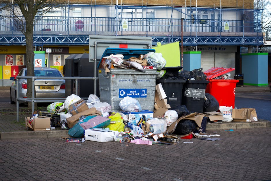 A collection of overflowing rubbish bins and scattered waste on a paved urban sidewalk outside a commercial building. The scene includes a large grey mixed paper and cardboard recycling bin with its lid partially open, revealing crumpled paper, cardboard boxes, and packaging waste. To the right, there are black and red waste containers, some bags overflowing with garbage, including plastic and paper waste. Additional rubbish such as flattened cardboard boxes, plastic bags, and miscellaneous packaging materials are strewn around the bins, spilling onto the surrounding paving stones. In the background, a metal railing separates the rubbish area from parked cars and a multi-storey building with blue scaffolding or protective netting, indicating ongoing construction or renovation. The environment appears to be a busy commercial or shopping area, with nearby storefronts and signage visible behind the rubbish. The scene highlights the importance of proper waste management and the challenges of private rubbish disposal outside of council collection services, suggesting a context suitable for independent waste removal solutions offered by services like Rubbish Removal Chiswick.