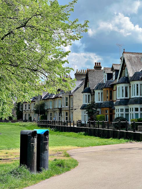 A row of Victorian-style terraced houses with bay windows and decorative brickwork lining a residential street. In the foreground, there are two black public rubbish bins positioned on a grassy verge next to a paved footpath. A large leafy tree with vibrant green foliage extends across the upper left corner, partially shading the scene. The background features a partly cloudy sky with patches of blue visible, and the environment appears clean and well-maintained, indicative of a typical suburban area where private rubbish collection and waste management services, such as those offered by Rubbish Removal Chiswick, support local residential waste removal needs.